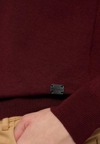 Bordový svetr s texturovaným pleteným designem, žebrovanými manžetami a černou značkovou nášivkou poblíž lemu. Ruka položená na béžovém oděvu.