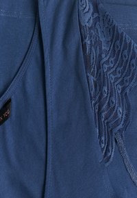 Modrý top bez rukávů vyrobený z měkkého materiálu s krajkovými detaily na jedné straně, kterému dominují květinové vzory a hladká textura. Obsahuje značku péče.