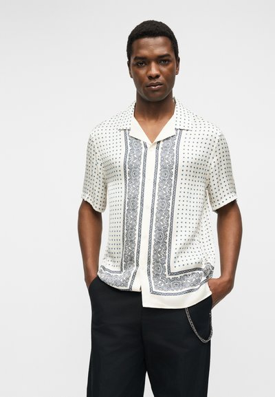 Άνδρας που φοράει λευκό κοντομάνικο πουκάμισο με μαύρα σχέδια πεϊζλί και σταυρούς, μαύρο παντελόνι με ασημένια αλυσίδα, στέκεται μπροστά σε απλό φόντο.