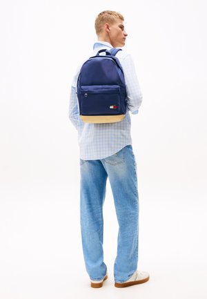 Mochila de tela navy con un fondo beige, bolsillo frontal con cremallera y acento de marca. Lleva puesta una camisa a cuadros azul claro y denim.