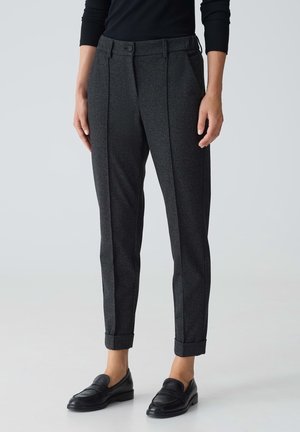 Femme portant un pantalon gris foncé sur mesure, longueur cheville avec revers et chaussures noires sans lacets, debout devant un fond neutre.
