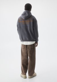 Grijze hoodie met oranje tekst en ontwerp op de achterkant, gecombineerd met loszittende, bruine broek en beige sneakers. Zachte, casual stof.