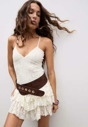 Robe en dentelle blanche avec des couches froncées, agrémentée d'une large ceinture en daim marron dotée de quincaillerie circulaire en or. Les bretelles spaghetti complètent le look.