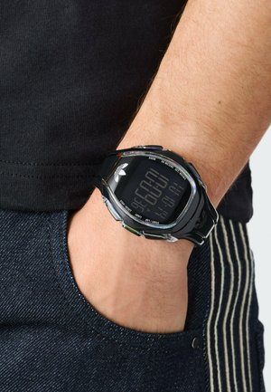 Skaitmeninis laikrodis su juodu dirželiu ir korpusu, stačiakampio formos ciferblatu. Ekrane rodomas laikmatis, o šonuose yra mygtukai funkcijoms.