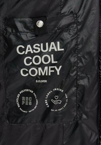 Sectie van zwarte nylon jas met een zak waarop in witte tekst staat: "CASHUAL COOL COMFY S.OLIVER." Cirkelvormige symbolen voor verzorging en materiaal eronder.