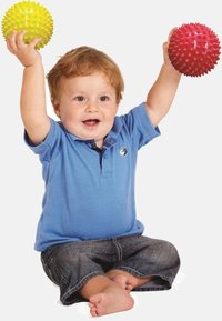 Ett litet barn sitter barfota, iklädd en blå skjorta och jeans, och håller glatt upp gula och röda taggiga sensoriska bollar över huvudet.