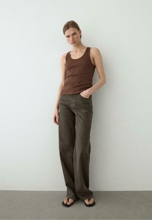Woman wearing a brown tank top, wide-leg dark brown pants, and black flip-flops, standing against a plain light wall.