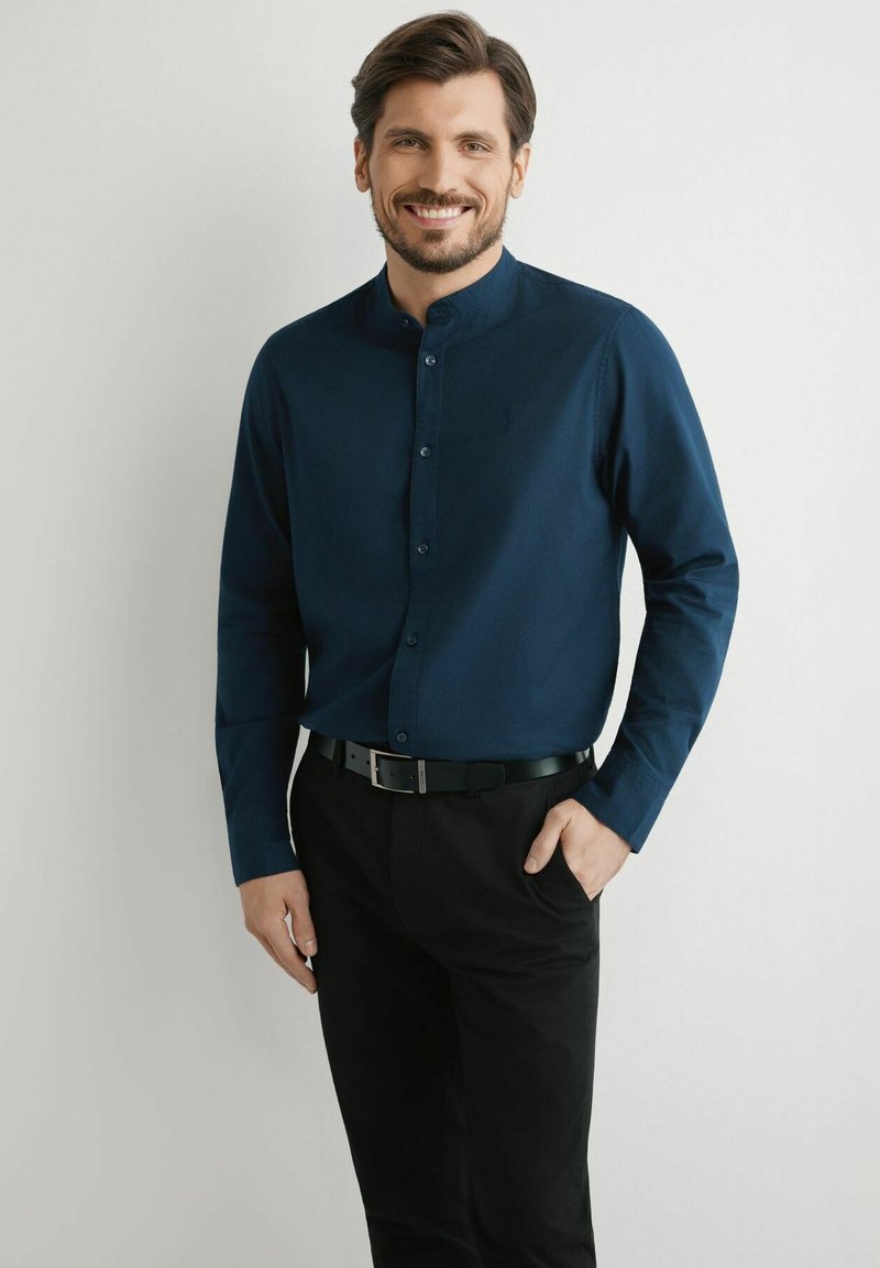 Uśmiechnięty mężczyzna z brodą, ubrany w zapinaną na guziki ciemnoniebieską koszulę i czarne spodnie, stojący z jedną ręką w kieszeni na jednolitym tle.