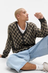 Cardigan în carouri maro și bej, cu nasturi în față. Purtați peste un tricou alb, asortat cu blugi evazați de culoare albastru deschis și adidași albi.