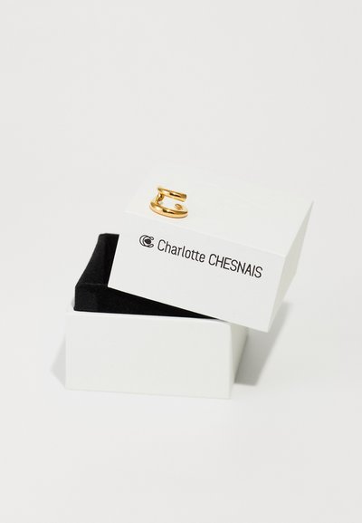 Inel din metal auriu într-un design sculptural, expus într-o cutie de bijuterii albă cu interior negru, care poartă numele mărcii Charlotte Chesnais.