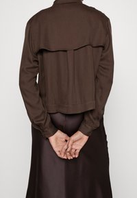 Veste courte marron en tissu lisse avec un rabat au dos, portée sur une longue jupe sombre, les mains jointes derrière le dos.