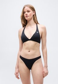 Νεαρή γυναίκα με μακριά ξανθά μαλλιά, που φοράει μαύρο μπικίνι με λουράκι στον λαιμό και ταιριαστά σλιπ, πάνω σε απλό ανοικτόχρωμο φόντο.