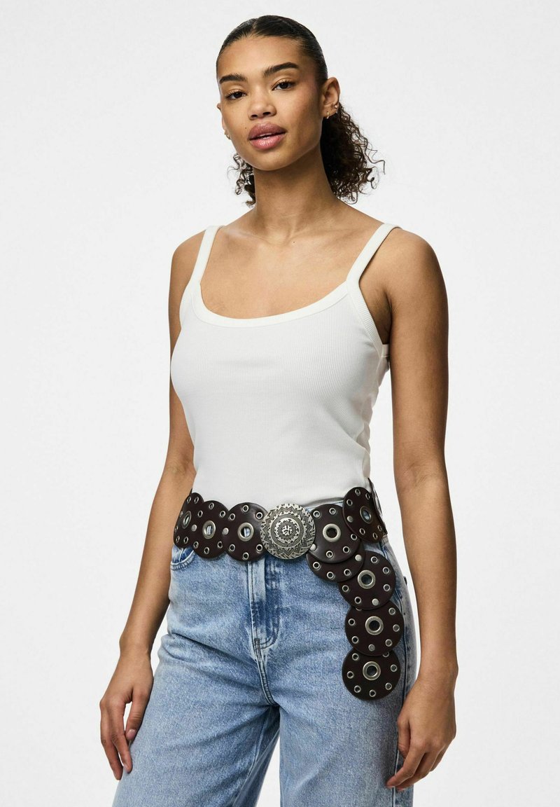 Bruine leren riem met een cirkelvormig uitgesneden ontwerp, zilveren metalen details, gedragen over een witte tanktop en blauwe spijkerbroeken.