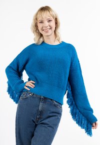 Kék kötött pulóver hosszú, bojtós ujjakkal. Texturált bordás mintázattal és kerek nyakkivágással rendelkezik, amelyet magas derékú farmernadrággal párosítottak.