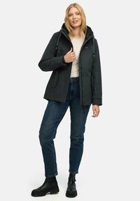 Nainen, joka pukee tummanharmaan hupparin, valkoisen paidan, siniset farkut ja mustat saappaat, seisoo käsi hupun vetskaria pitkin.