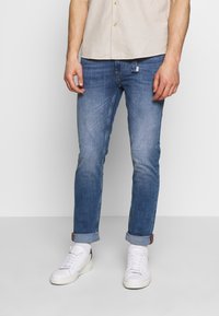 Mörkblå denimjeans med en ljus tvätt, uppvikta fållar och ett märke på baksidan. Matchas med vita sneakers och en beige knappad skjorta.