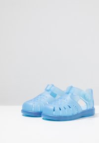 Heledad sinised läbipaistvad sandaalid, suletud varbaga ja lõigatud disainiga. Pehme plastmaterjal, reguleeritav rihm ja sinine gradient talla.