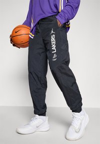 Person i sorte Los Angeles Lakers bukser, lilla langærmet top, holder en basketball, iført hvide Nike sneakers og hvide sokker.