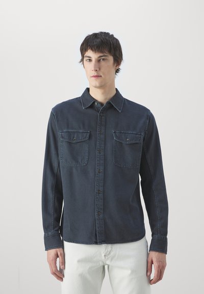 Mørkeblå denimskjorte med knaplukning, spredt krave og to brystlommer. Har lange ærmer og en afslappet pasform.
