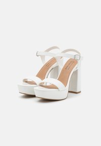 White platform sandals with a thick block heel, open toe, and ankle strap featuring a silver buckle. Smooth texture, minimalist design.
