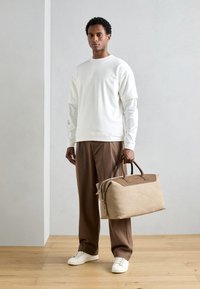 Beige taška na cesty s hnědými koženými akcenty, strukturovaný povrch a dvě ucha. Modelka v bílém topu a hnědých kalhotách stojí na dřevěné podlaze.