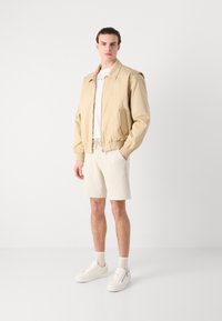 Giubbotto bomber beige con polsini e vita elasticizzati, indossato sopra una maglietta bianca, abbinato a pantaloni corti chiari e sneakers bianche.