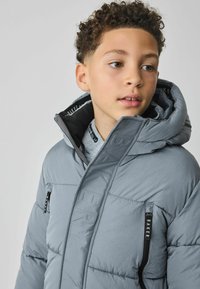 Doudoune gris clair avec une poche zippée et un logo visible. Dotée d'un col haut et d'une capuche, fabriquée en matériau soyeux.