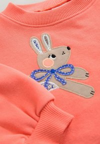 Sweatshirt corail avec design de lapin brodé. Le lapin porte un nœud papillon à pois bleu et des accents beiges. Manches et col côtelés.