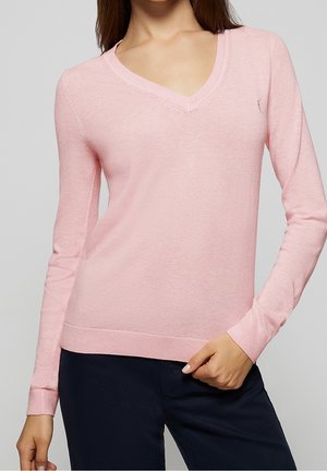 Femme portant un pull à manches longues rose clair avec un col en V et un pantalon foncé, debout avec une main reposant près de la taille.