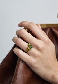 Dzeltens metāla gredzens ar diviem akmeņiem: zaļu apaļu un sirds formas dzidru akmeni. Roka tur brūnu teksturētu ādas somu.