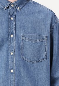 Camasa din denim, cu nasturi, de culoare albastru deschis. Prezintă un guler ascuțit, un buzunar frontal și nasturi albi. Textură moale, cu un croi clasic.