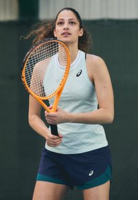 Tennis speler houdt een oranje racket vast, gekleed in een lichtblauw, mouwloos topje met een ruitpatroon en donkerblauwe shorts met een groene accent.