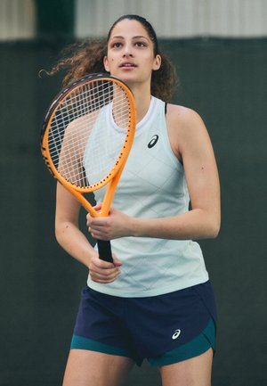 Un joueur de tennis tient une raquette orange, portant un haut sans manches bleu clair avec un motif en losanges et un short bleu foncé avec une touche de vert.