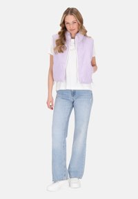 Gilet imbottito lavanda con colletto rialzato, chiusura con zip, tasche laterali; indossato sopra una camicia bianca e abbinato a jeans azzurri e sneaker bianche.