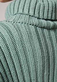 Sweter z golfem w miękkim zielonym kolorze, z pionowym żebrowanym wzorem. Szczegóły obejmują luźny krój i przytulny, podwinięty kołnierz.