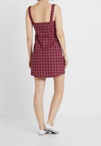 Abito a quadri rosso e nero con spalline larghe, schiena quadrata e vita aderente, abbinato a sneakers bianche. Tessuto dalla texture liscia.