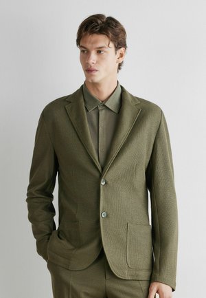 Junger Mann in einem olivgrünen Anzug mit einer zugeknöpften Jacke und einem Hemd mit Kragen, die Hände in den Taschen, leicht nach links gewandt vor einem schlichten Hintergrund.