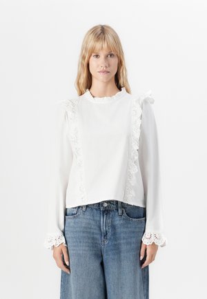 Blandet kvinne med blondt hår iført en hvit langermet bluse med blondedetaljer og vide blå jeans, stående mot en ensfarget hvit bakgrunn.