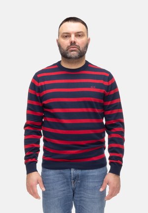 Uomo con capelli corti e scuri e barba, indossa un maglione blu navy con strisce orizzontali rosse e jeans blu, in piedi davanti a uno sfondo bianco.