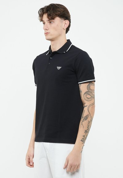 Jonge man met golvend haar, gekleed in een zwart poloshirt met witte biezen en witte broek, toont een slangentattoo op zijn rechterarm.