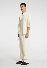 Jasnobeżowa koszula z krótkim rękawem i kołnierzykiem w połączeniu z pasującymi, luźnymi spodniami. Czarna obuwie dopełnia strój, charakteryzujące się teksturowanym wykończeniem materiału.