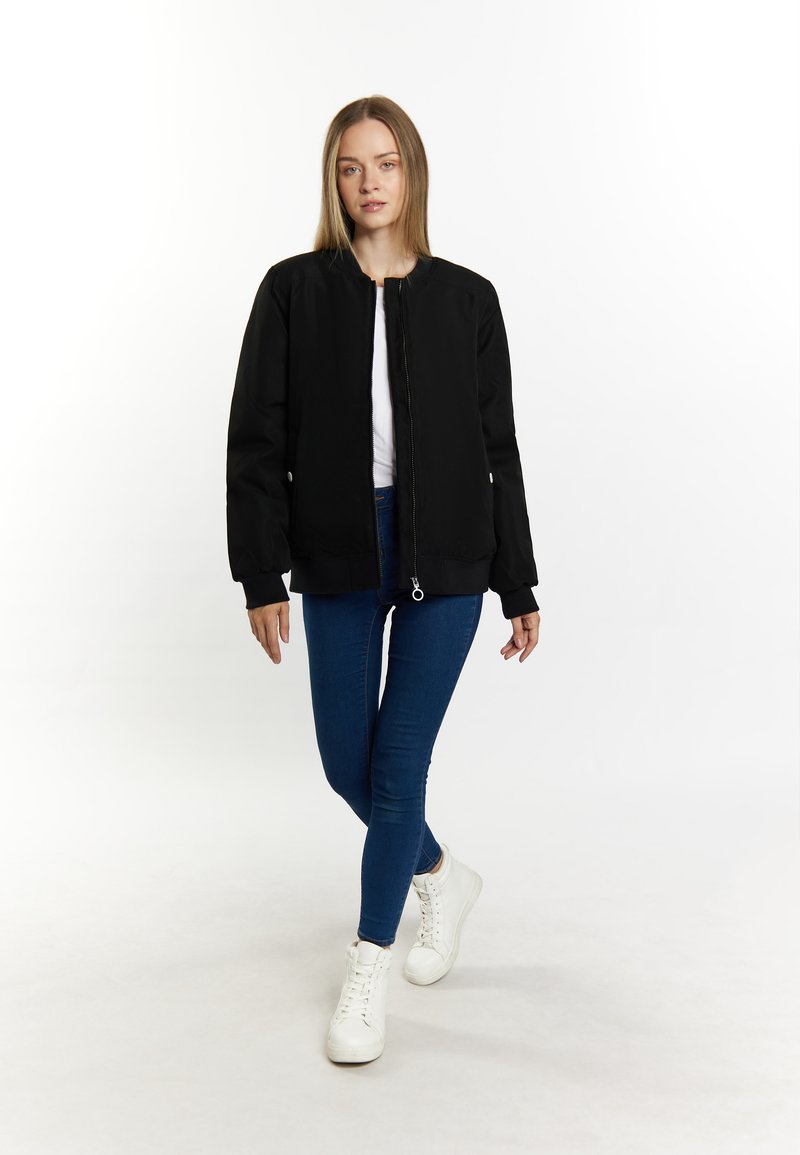 Schwarze Bomberjacke mit einem Frontreißverschluss, gerippten Bündchen und Saum. Kombiniert mit blauen Skinny-Jeans und weißen Turnschuhen. Modell geht.
