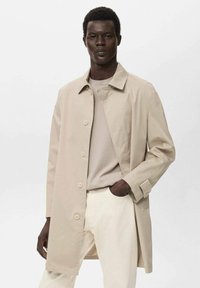 Beige Leichtjacke mit Kragen und sechs Knöpfen, kombiniert mit einem cremefarbenen Hemd und hellen Hosen, präsentiert an einem Modell.