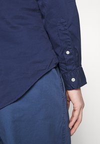 Námořnická modrá košile na knoflíky s hladkou texturou, s špičatým límečkem, dvouknotovými manžetami a zakřiveným lemem.