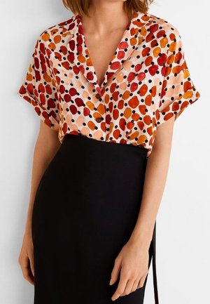 Femme portant un chemisier à manches courtes avec des taches abstraites rouges, orange et noires, rentrée dans une jupe noire taille haute.