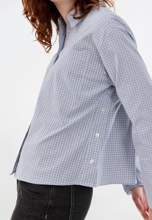 Femme portant une chemise boutonnée à carreaux bleu clair avec des boutons latéraux et un jean noir, vue de profil des épaules aux hanches.