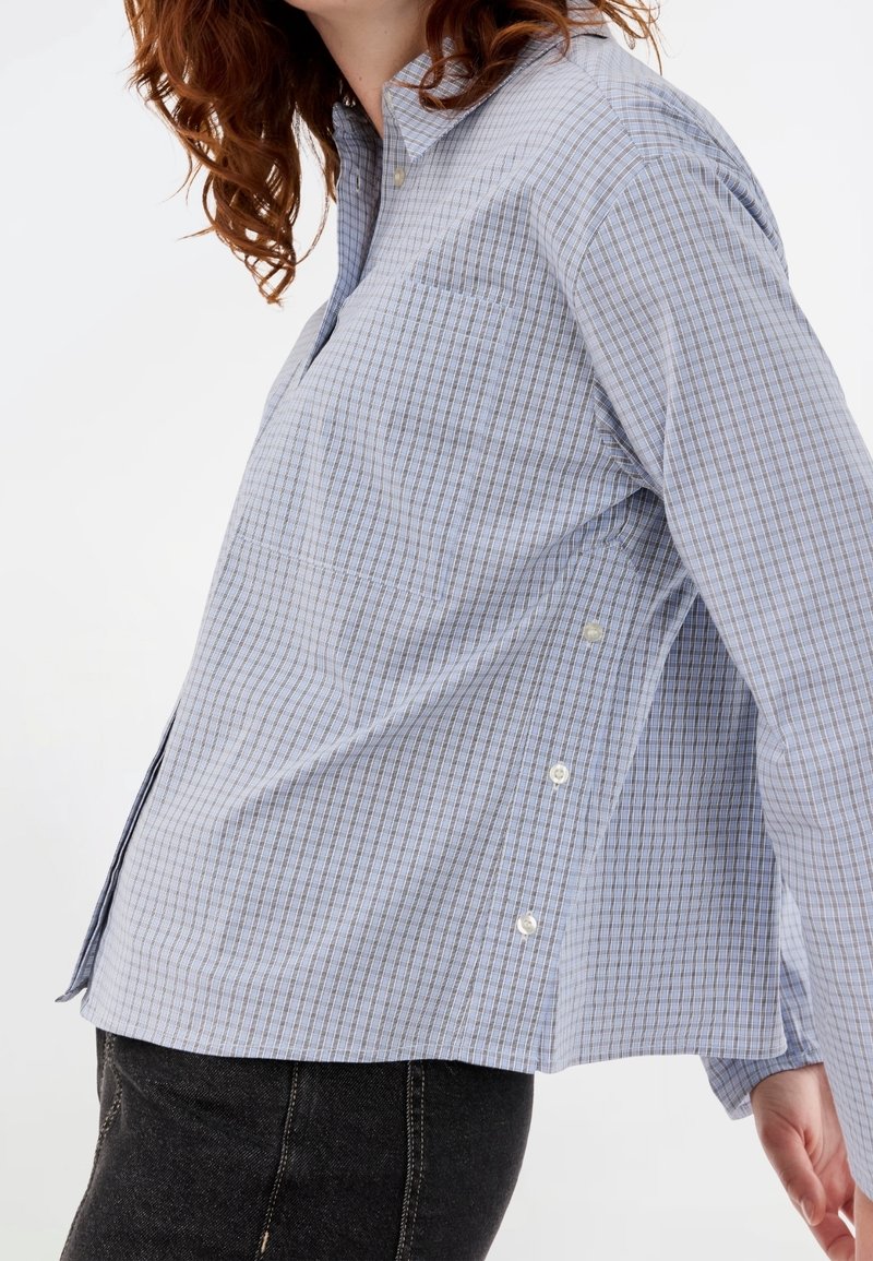 Femme portant une chemise boutonnée à carreaux bleu clair avec des boutons latéraux et un jean noir, vue de profil des épaules aux hanches.