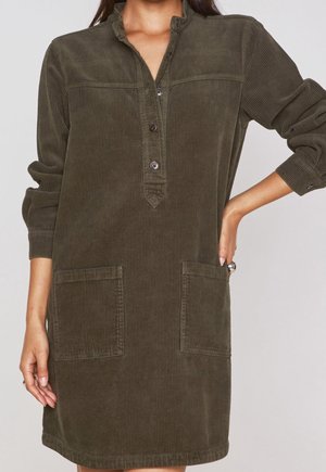 Kvinde bærer olivengrøn kordfløjls kjole med lange ærmer, knappet opslag og to forlommer, står med den ene hånd på hoften.
