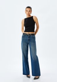 Zwarte mouwloze top gecombineerd met wijde lichtblauwe denim jeans. Zwarte riem met een ronde zilveren gesp. Punttop zwarte schoenen met zilveren accenten.