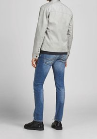 Homme portant une veste gris clair, un jean slim bleu et des chaussures noires, debout de dos sur un fond uni.